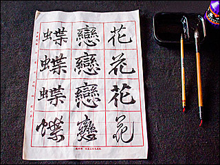 Three Chinese characters in four different calligraphy scripts written on calligraphy grid paper, with two brushes, a bottle of ink, and an inkstone at the side.