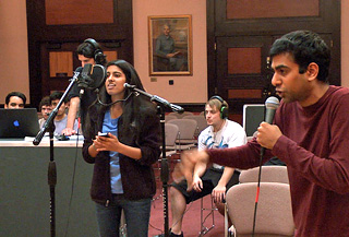 Photo of two singers on microphones, with a recording engineer standing in the background monitoring the session on a laptop.