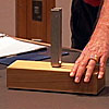 A tuning fork sits on a block of wood. The professor holds down one end with his hand.