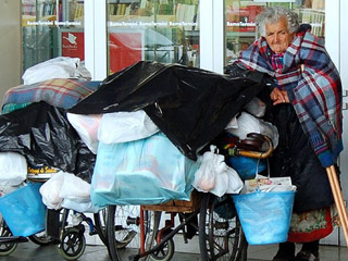 An elderly woman on crutches waits outside a train station with bags, buckets, and blankets resting on two wheelchairs beside her. An elderly woman on crutches waits outside a train station with bags, buckets, and blankets resting on two wheelchairs beside her.